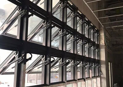 Eine Reihe von großen, schwarz gerahmten, nach außen zu öffnenden Fenstern mit mechanischen Armstützen ist entlang einer Wand im industriellen Stil installiert.