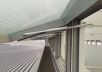 Eine Reihe von automatisierten Fensterantrieben, die an Glasfenstern installiert sind, mit Blick auf ein Metalldach und einen Industrieweg draußen.