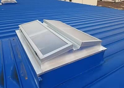 Ein Metalldach mit einer rechteckigen, hochgezogenen Oberlicht-Lüftungsstruktur mit gewinkelten, lichtdurchlässigen Paneelen und blauer Wellblecheindeckung.